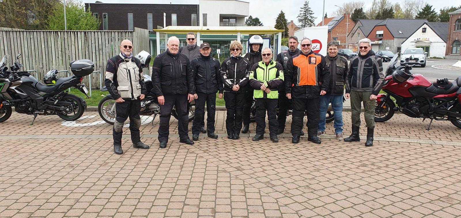 Een groep rijders poseert op een kasseiplein bij een bushalte, met motoren links en rechts.