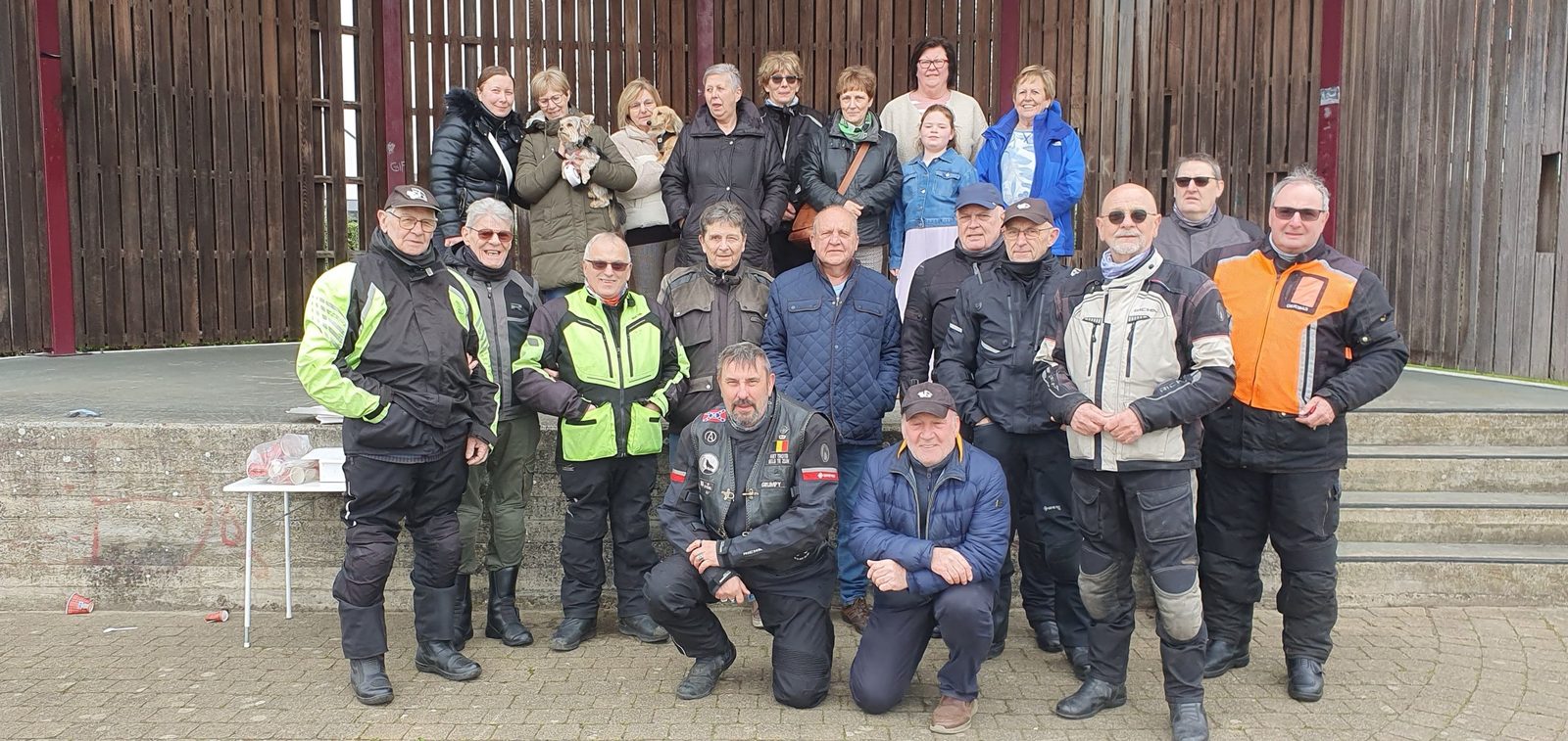 Een grote groep rijders en partners poseert voor een houten podium.