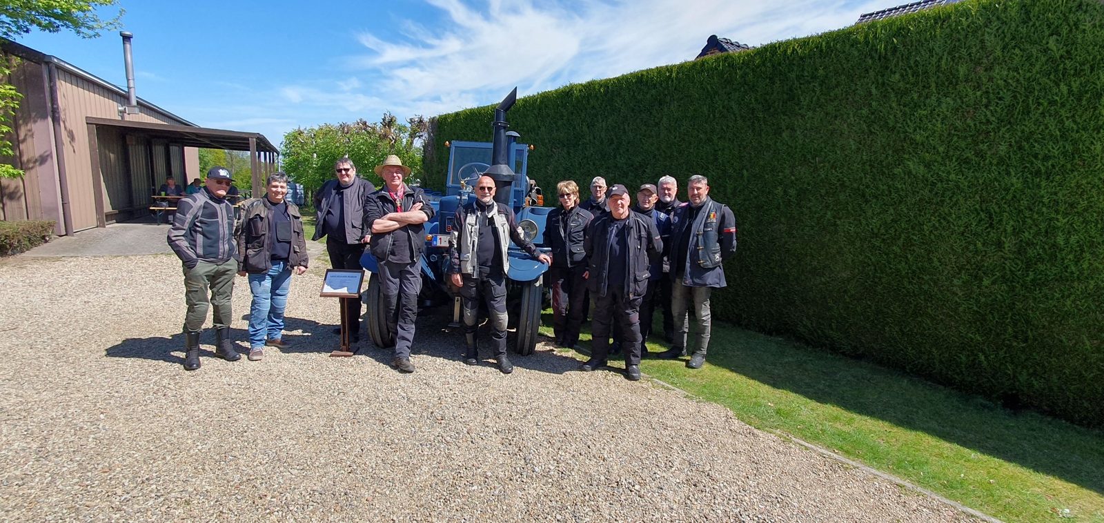 Een groep rijders poseert op een grindpad voor een blauwe tractor en een hoge haag.