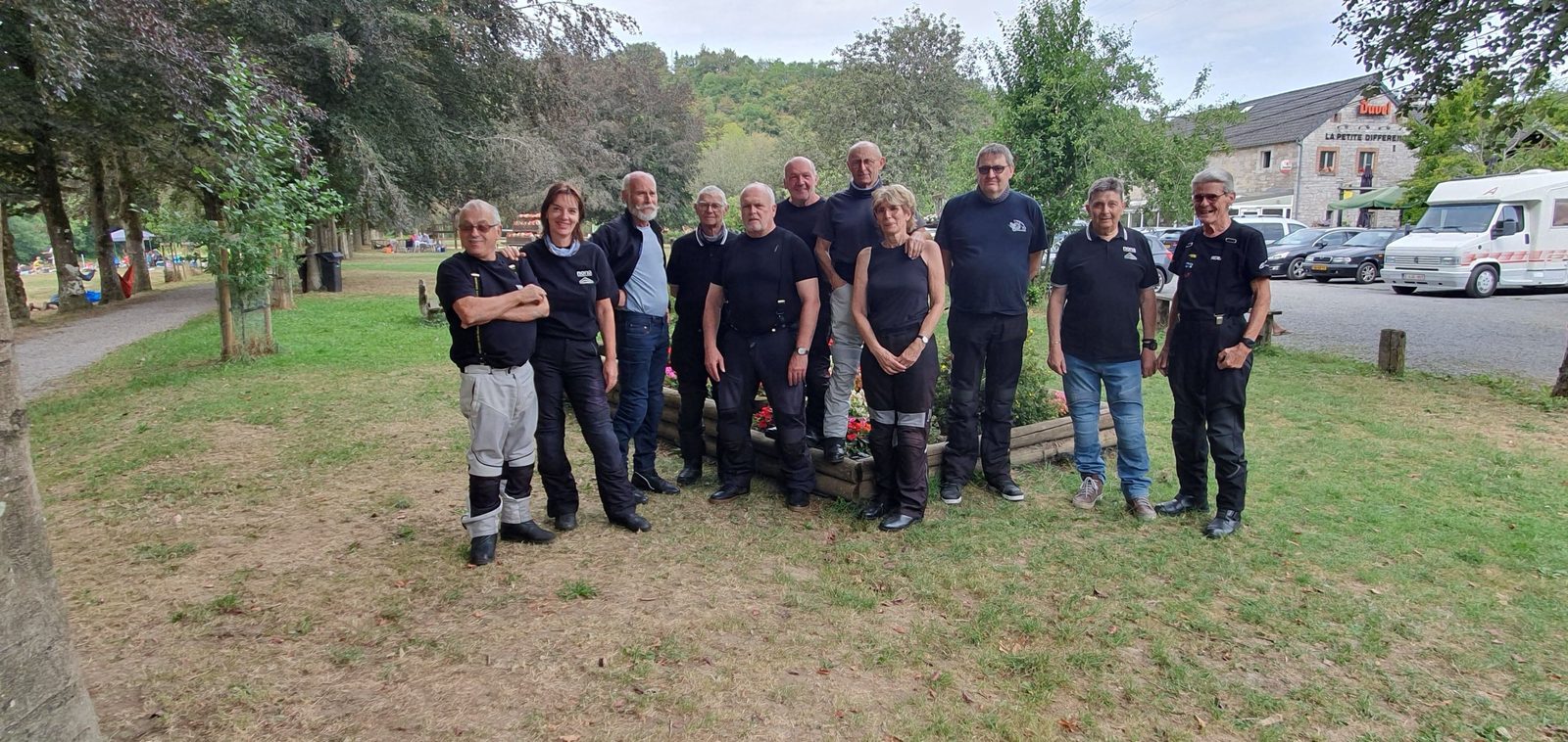Een groep rijders staat op het gras bij een bloembak, met een café op de achtergrond.