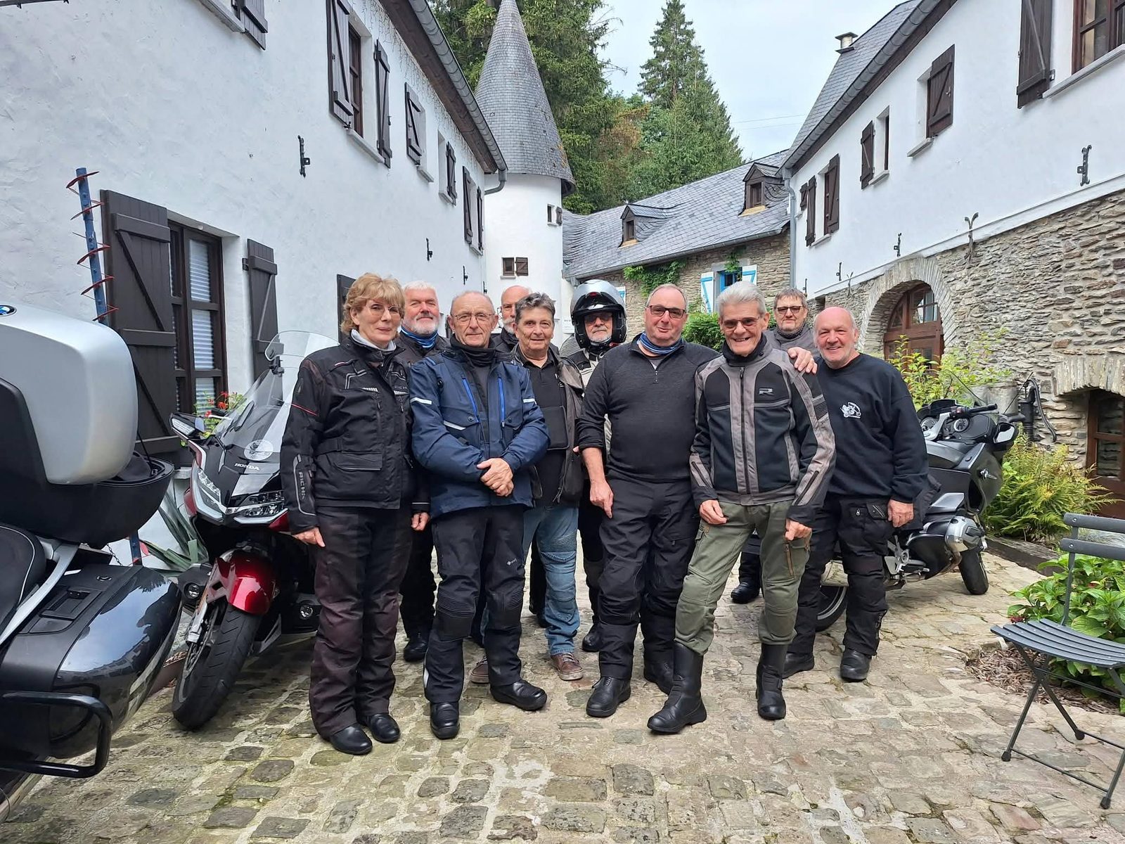 Een groep rijders poseert op een kasseiplein tussen witte huizen, met motoren op de achtergrond.